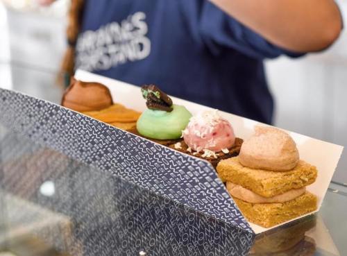 A variety of colorful desserts displayed in a box, including green, pink, and brown treats, with a person in the background.