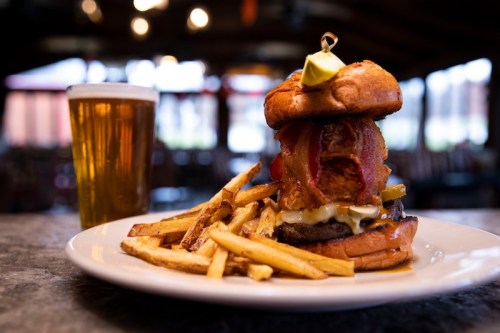 A towering burger with bacon, pickles, and cheese, served with fries and a glass of beer in a cozy restaurant setting.