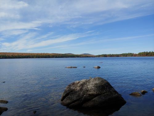 A serene lake scene with a large rock in the foreground, surrounded by calm water and distant hills under a blue sky.