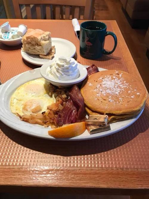 A breakfast plate with eggs, bacon, hash browns, pancakes topped with powdered sugar, and an orange slice. Coffee on the side.