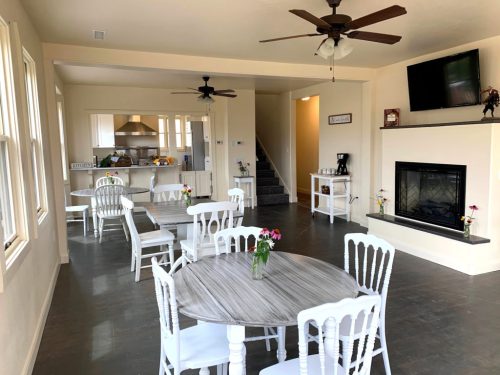 Bright, open living space with white chairs, tables, and a fireplace, featuring a kitchen and staircase in the background.