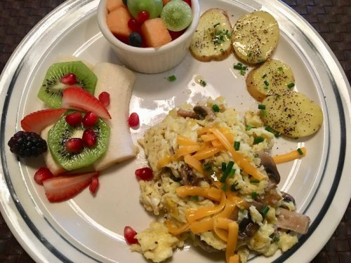 A plate with scrambled eggs topped with cheese, sautéed mushrooms, sliced potatoes, and a fruit medley of melon and berries.