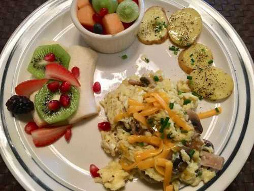 A plate with scrambled eggs topped with cheese, sautéed mushrooms, sliced potatoes, and a fruit medley of melon and berries.