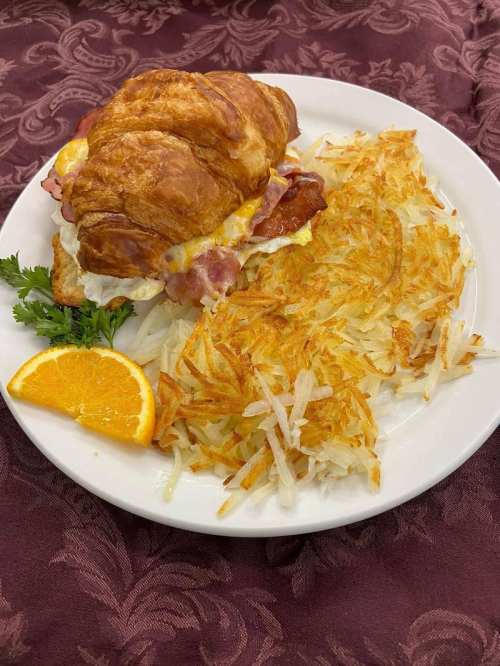 A croissant sandwich with bacon and cheese, served with crispy hash browns and a slice of orange on a white plate.