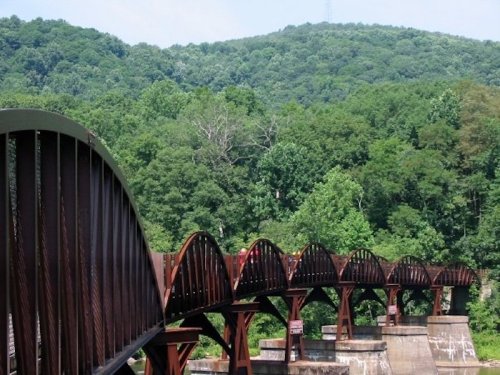 A curved metal bridge spans over a river, surrounded by lush green hills and trees under a clear blue sky.