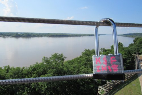 A padlock hangs on a railing overlooking a calm river and lush greenery under a clear blue sky.