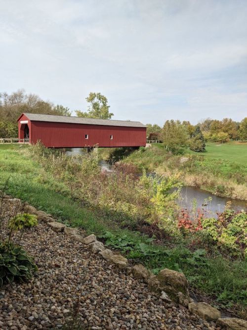 A red covered bridge spans a calm stream, surrounded by lush greenery and autumn foliage.