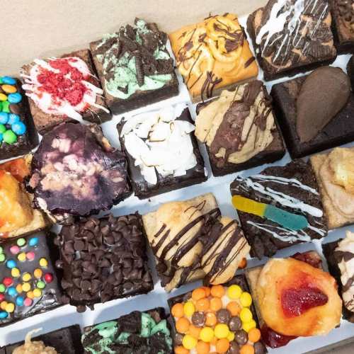 A variety of colorful brownies topped with different candies, frosting, and drizzles, arranged in a box.