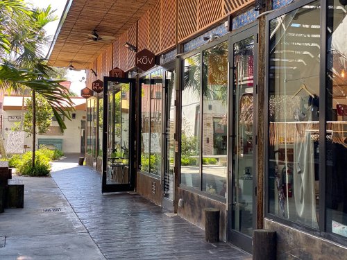A sunny outdoor shopping area with glass storefronts and a paved walkway lined with greenery.