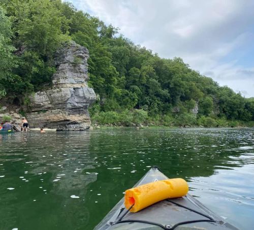 A kayak on calm water with a yellow foam float, surrounded by green trees and rocky cliffs in the background.
