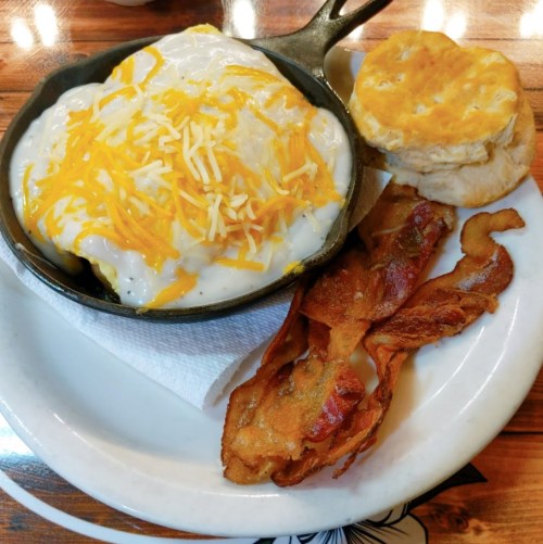 A skillet of creamy biscuits and gravy topped with cheese, served with crispy bacon and a biscuit on the side.