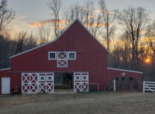 A red barn with white trim stands against a sunset, surrounded by trees and a grassy field.