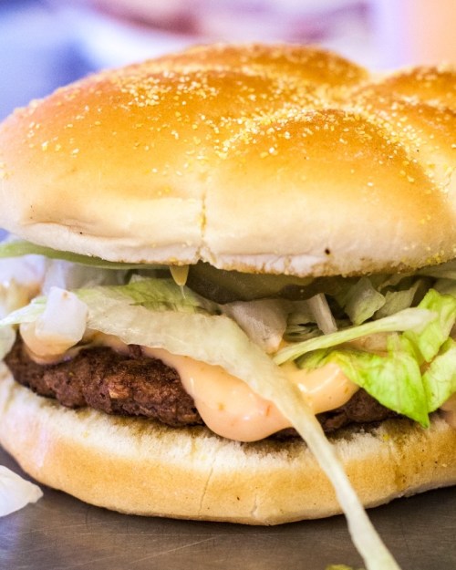 A close-up of a burger with a sesame seed bun, lettuce, and creamy sauce peeking out from the sides.