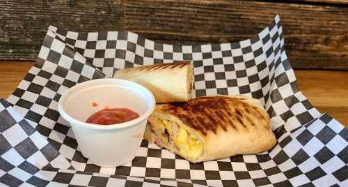 A grilled burrito cut in half on a checkered paper, served with a small cup of salsa.