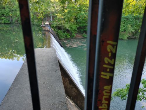 A view of a dam with water cascading over the edge, framed by metal railings and surrounded by lush greenery.