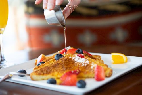 A hand pours syrup over a plate of French toast topped with berries and orange slices, with a glass of juice in the background.