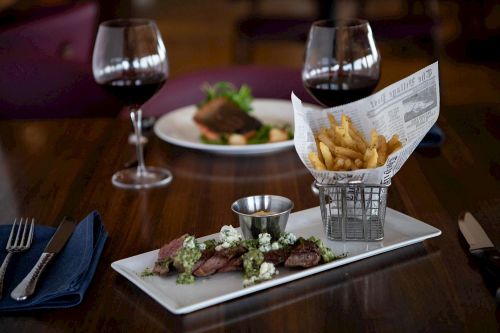 A plate with sliced steak topped with green sauce and blue cheese, served with fries in a basket and red wine glasses.
