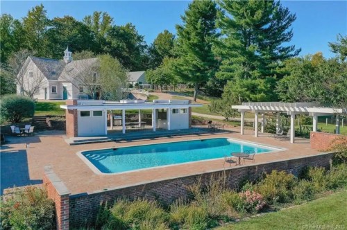 Aerial view of a pool area with a patio, surrounded by greenery and a house in the background. Clear blue sky above.