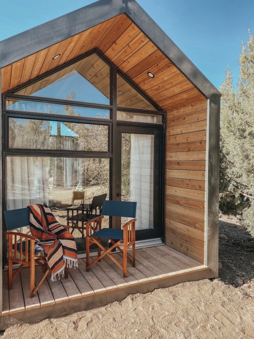 A modern wooden cabin with large windows, featuring two chairs and a cozy blanket on the porch.