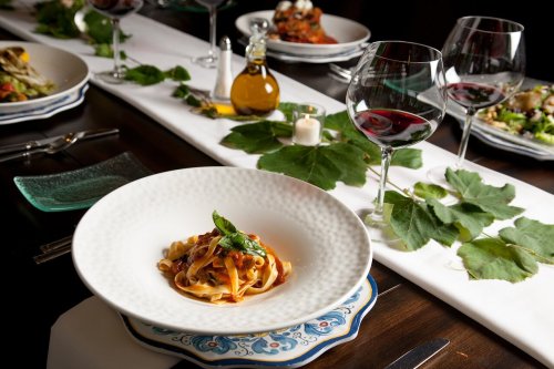 A beautifully set dining table featuring a plate of pasta, wine glasses, and various dishes surrounded by green leaves.