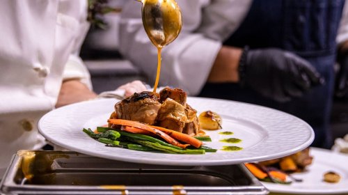 A chef drizzles sauce over a beautifully plated dish featuring meat, carrots, and green vegetables.