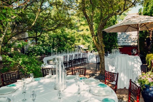 Outdoor dining setup with tables, chairs, and umbrellas surrounded by greenery on a sunny day.