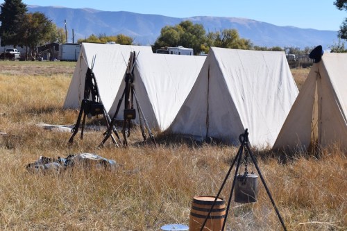 Canvas tents set up in a grassy field, with mountains in the background and cooking equipment nearby.
