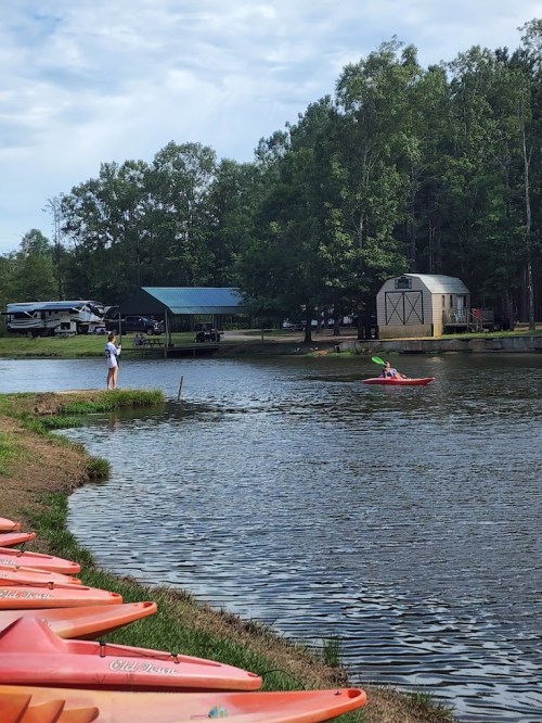 A serene lake scene with kayaks lined up, a person fishing, and another kayaking in the background among trees.