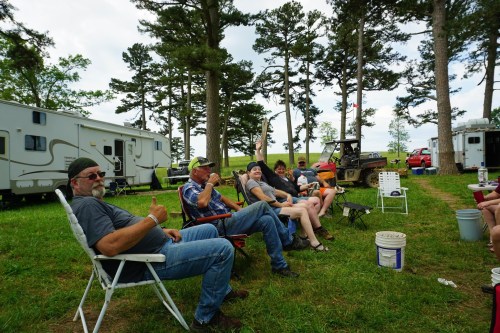 A group of people sitting in lawn chairs outdoors, enjoying a gathering near RVs and trees.