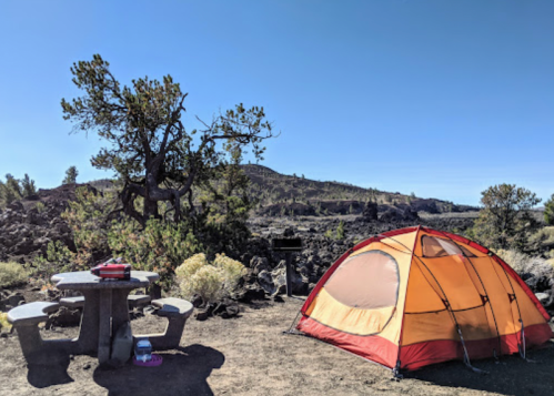A bright orange tent set up near a picnic table in a rocky campsite with trees and hills in the background.