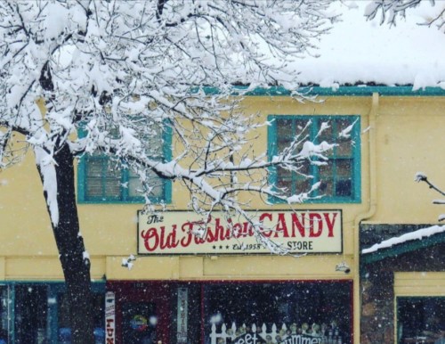 A quaint candy store sign, "The Old Fashioned Candy," surrounded by snow-covered trees and a wintery scene.