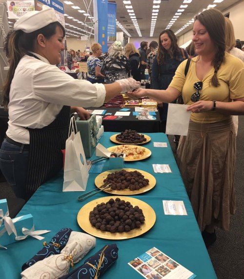 A vendor hands a sample to a customer at a food fair, with trays of chocolate truffles on the table.