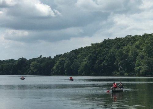 Two canoes with people paddling on a calm lake surrounded by lush green trees under a cloudy sky.