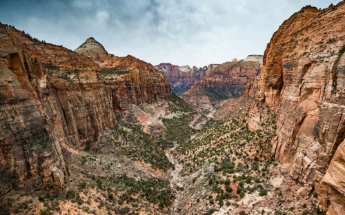 A panoramic view of a rugged canyon with steep red rock walls and a winding river below, under a cloudy sky.
