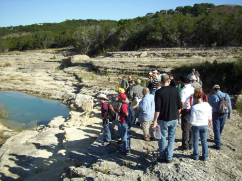 A group of people gathered near a rocky area by a small body of water, surrounded by trees and greenery.