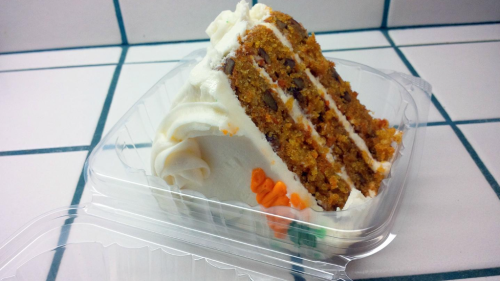 A slice of carrot cake with cream cheese frosting in a clear plastic container, decorated with orange accents.