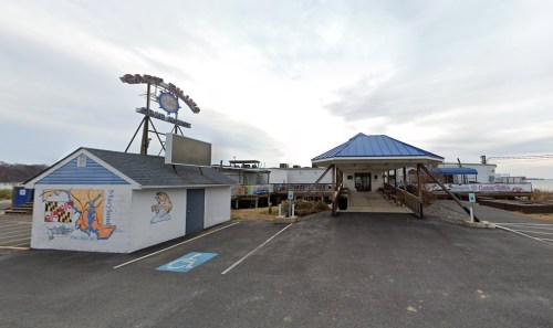 A waterfront restaurant with a blue roof and outdoor seating, featuring a sign and nearby parking area.