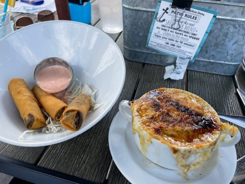 A bowl of spring rolls with dipping sauce and a dish of French onion soup, both served on a wooden table.