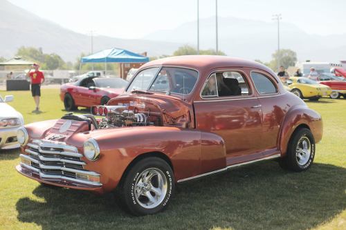 A classic orange hot rod car with a shiny engine on display at a car show, set on a grassy field.