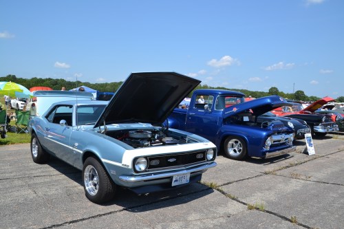 Two classic cars are parked side by side at a car show, with hoods open under a clear blue sky.