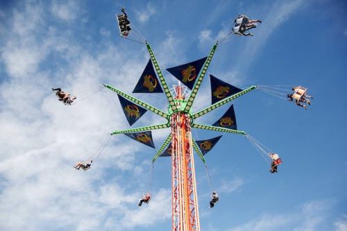 A colorful swing ride with people soaring high against a blue sky, surrounded by clouds and vibrant flags.
