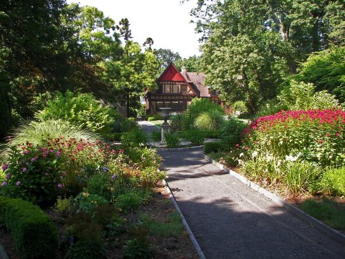 A scenic garden path lined with colorful flowers, leading to a charming house surrounded by lush greenery.