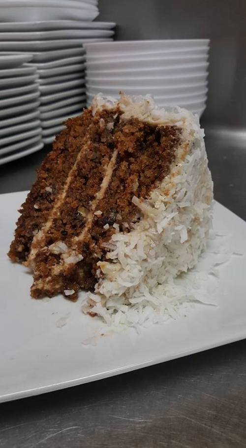 A slice of coconut cake with layers of brown cake and white frosting, garnished with shredded coconut on a white plate.