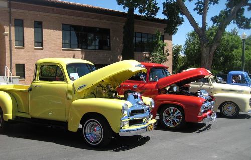 A row of classic trucks with hoods open, showcasing their engines, parked in front of a brick building.