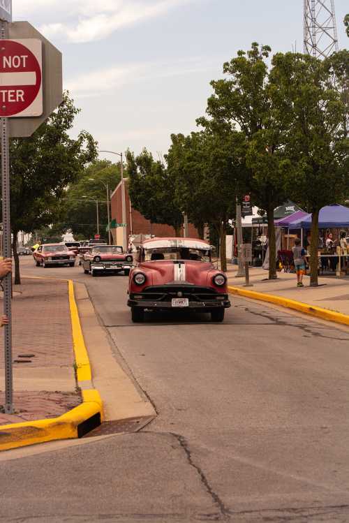 A vintage red car drives down a tree-lined street, with other classic cars in the background and a stop sign nearby.