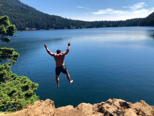 A man jumps off a rocky ledge into a calm lake surrounded by trees and mountains on a sunny day.