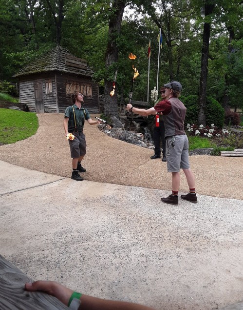 Two performers juggle flaming torches in a park setting, with a wooden building and trees in the background.