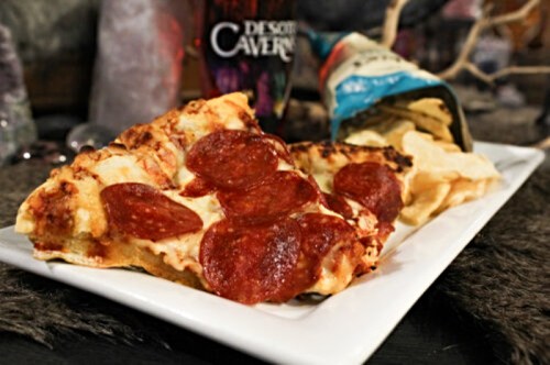 A slice of pepperoni pizza on a white plate next to a bag of chips, set on a textured surface.