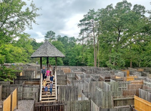 Aerial view of a wooden maze with people walking on a platform, surrounded by trees and cloudy skies.