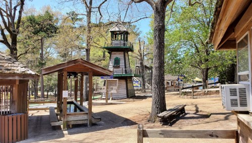 A playground area featuring a tall watchtower, wooden structures, and picnic tables surrounded by trees.